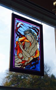 A stained glass picture of two people embracing, in a wooden frame hung in front of a large window.