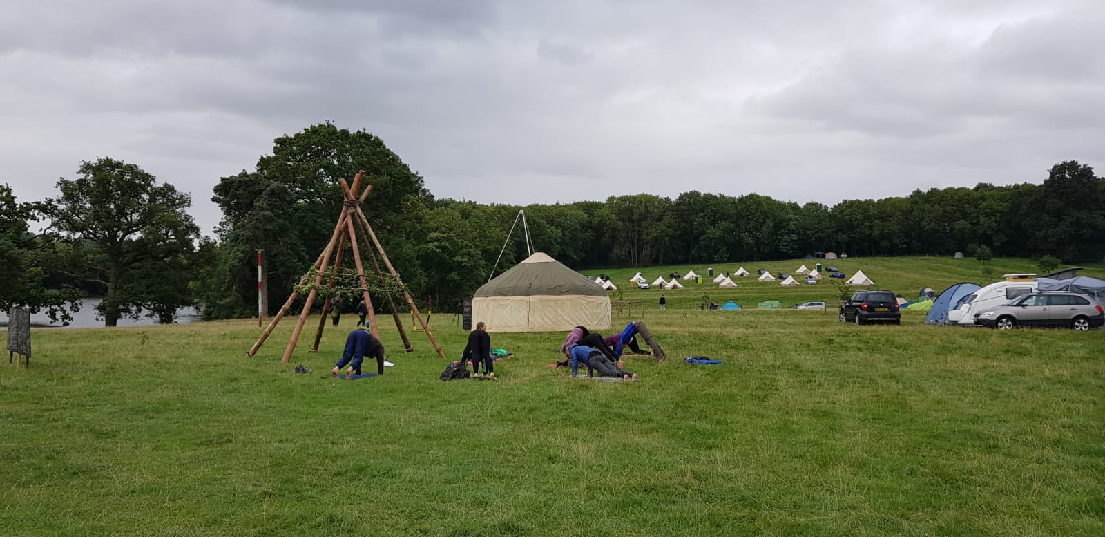 A large green field with a small group of people doing yoga. In the back ground white tents socially distanced.