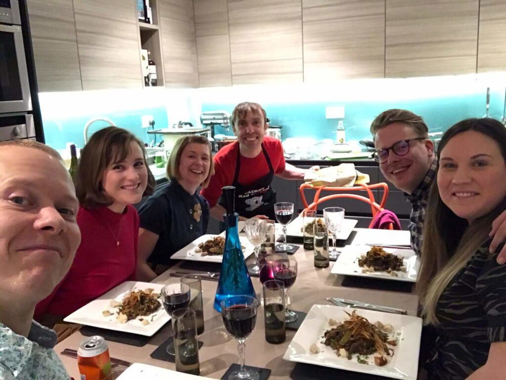 6 people sat around a dining table with food and drinks. A kitchen in the background.