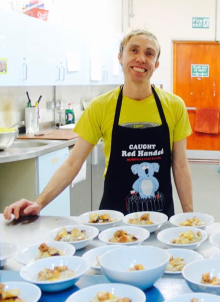 A man standing in a kitchen wearing an apron. Lots of bowls of food in front of him.